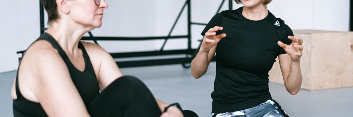 Two women in sportswear sitting on a yoga mat, having a detailed discussion in a gym.